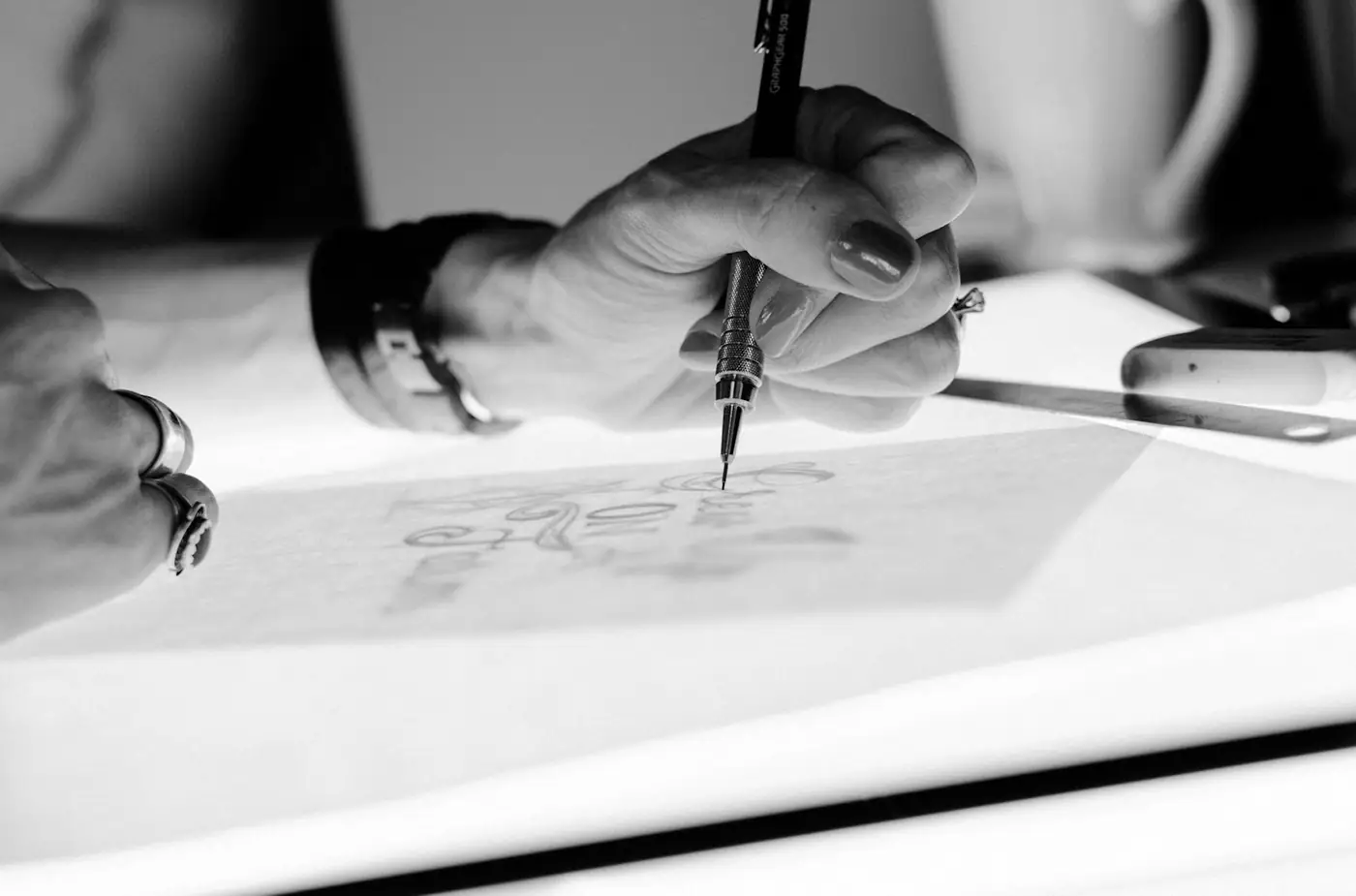 A hand holding a pen over a drawing on a light table, the sheet glowing from below