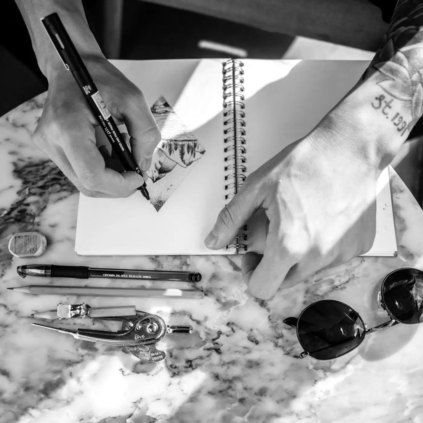 Two hands inking a drawing in a sketchbook, pens and a compass spread across the desk